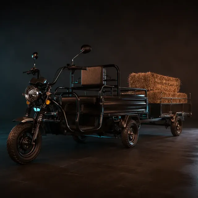 Three-wheeled electric cargo tricycle towing a flatbed trailer loaded with hay bales on a farm road at golden hour
